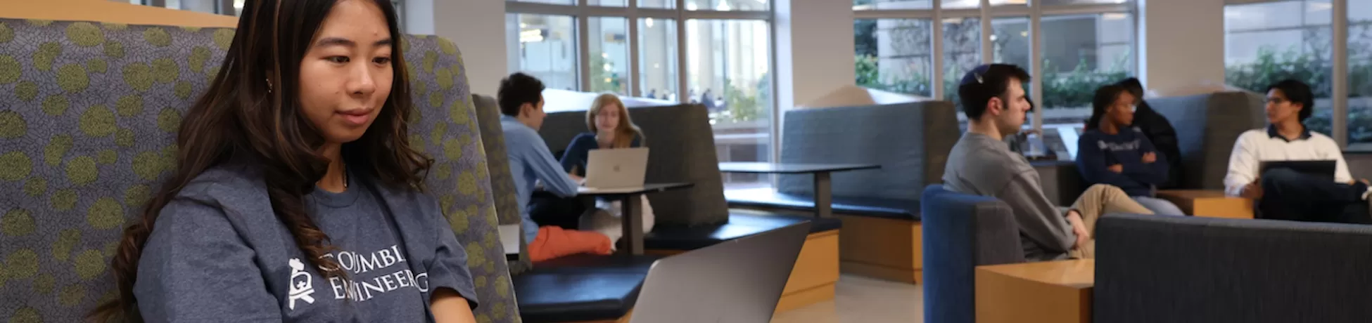 close up photo of girl in columbia engineering shirt using laptop at a booth, background with other groups of students sitting together