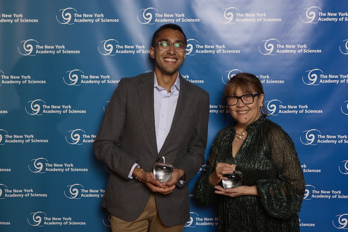  Two people smile while holding glass awards in front of a blue step-and-repeat backdrop reading “The New York Academy of Sciences.”